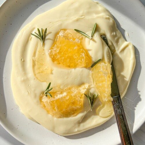 A white plate with creamy yogurt and whipped Brie, topped with honeycomb pieces and rosemary sprigs; a butter knife rests on the plate.