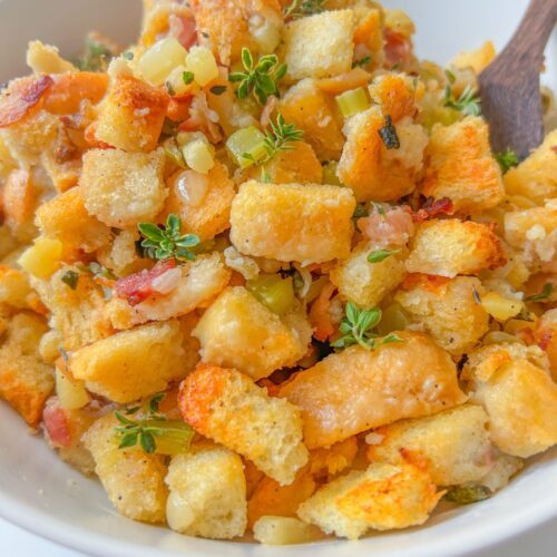 A bowl of homemade stuffing made with golden toasted bread cubes, diced celery, herbs, and small bits of bacon, garnished with fresh thyme—this is truly the Best Stuffing Recipe, with a wooden spoon partially visible.