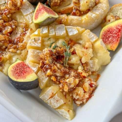 A close-up of Baked Hazelnut Brie topped with chopped nuts and honey, garnished with fresh figs and a sprig of thyme, served in a white dish.