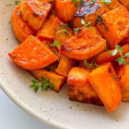 A speckled ceramic plate filled with Roasted Sweet Potatoes, garnished with fresh green herbs. The sweet potatoes are golden, slightly caramelized, and bursting with savory-sweet flavor.
