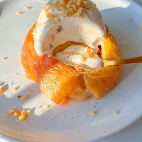A classic dessert featuring baked apples topped with a scoop of vanilla ice cream and crushed nuts, elegantly served on a white plate with a spoon scooping into the warm, sweet treat.