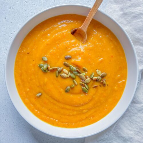 A bowl of creamy Roasted Butternut Squash Soup topped with pumpkin seeds, with a wooden spoon resting inside. The bowl sits on a light surface next to a textured white cloth.