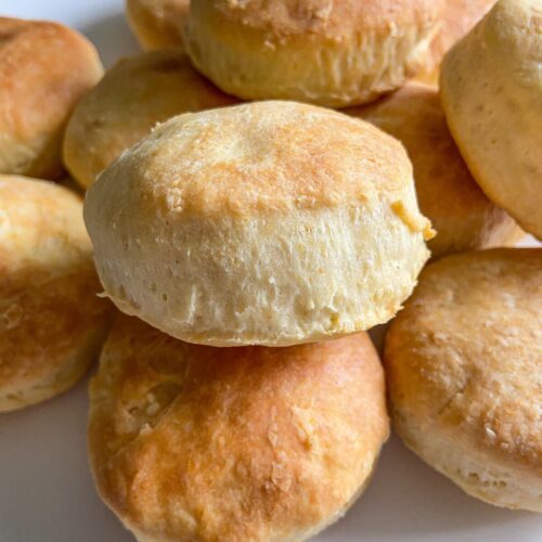 A close-up of a plate stacked with golden brown, freshly baked buttermilk biscuits on a white background. The biscuits have a soft, fluffy texture with lightly crisp tops.