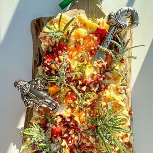 A festive Holiday Butter Board topped with nuts, fruit preserves, herbs, and sugared rosemary sprigs, arranged on a wooden board with two decorative cheese knives and a green ribbon bow.