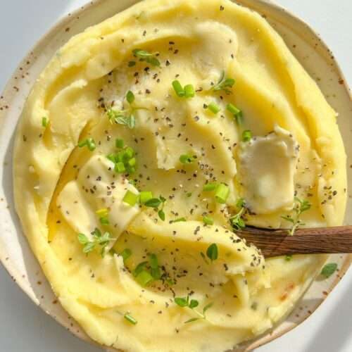 A plate of creamy Roasted Garlic Mashed Potatoes topped with butter, cracked black pepper, and fresh green herbs, with a wooden spoon resting in the dish.