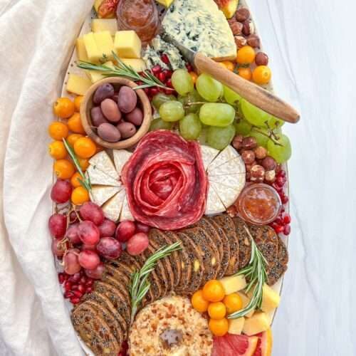 A Holiday Cheese Board featuring various cheeses, salami roses, fresh figs, grapes, cherry tomatoes, olives, crackers, grapefruit slices, rosemary sprigs, and bowls of jam is artfully arranged on a white surface with a linen cloth.