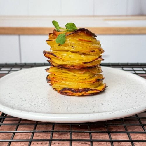 A neatly stacked tower of greek lemon potato stacks, thinly sliced and roasted, garnished with a small green herb, served on a white plate with a speckled pattern, placed on a black cooling rack.