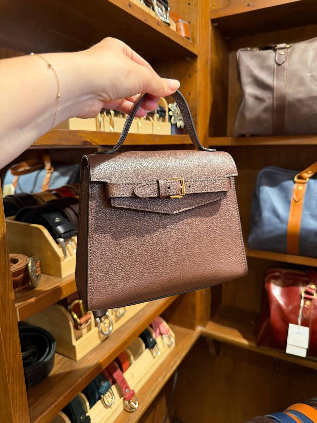 A hand holds a small brown leather handbag with a top handle and buckle, displayed in a store with shelves of belts and bags in the background.