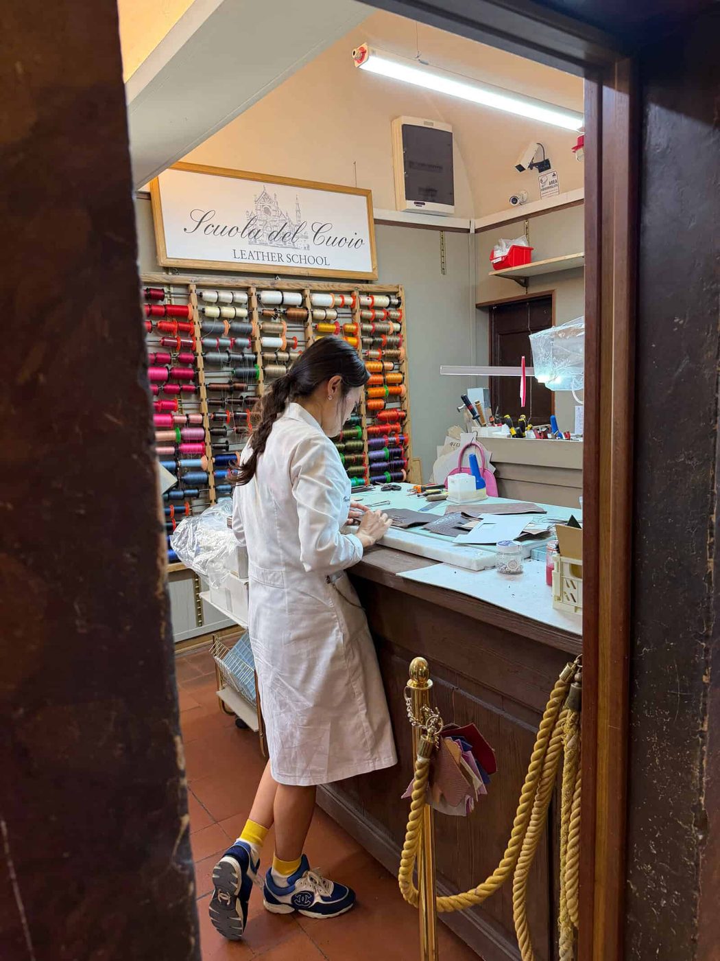 A person in a white lab coat works at a counter in a leather school, with colorful threads organized on the wall behind them and various leatherworking tools on the desk.