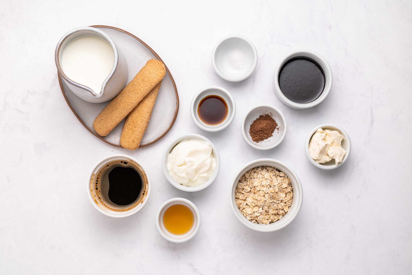 Assorted ingredients in small bowls on a white surface, including milk, ladyfinger biscuits, coffee, oats, yogurt, cocoa powder, vanilla, syrup, mascarpone cheese, salt, and sweetener.