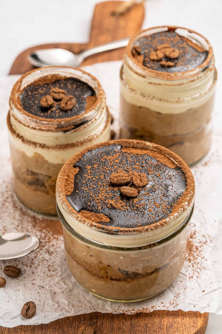 Three glass jars filled with layered tiramisu dessert, topped with cocoa powder, chocolate, and coffee beans, are arranged on a wooden board with a spoon and scattered coffee beans nearby.