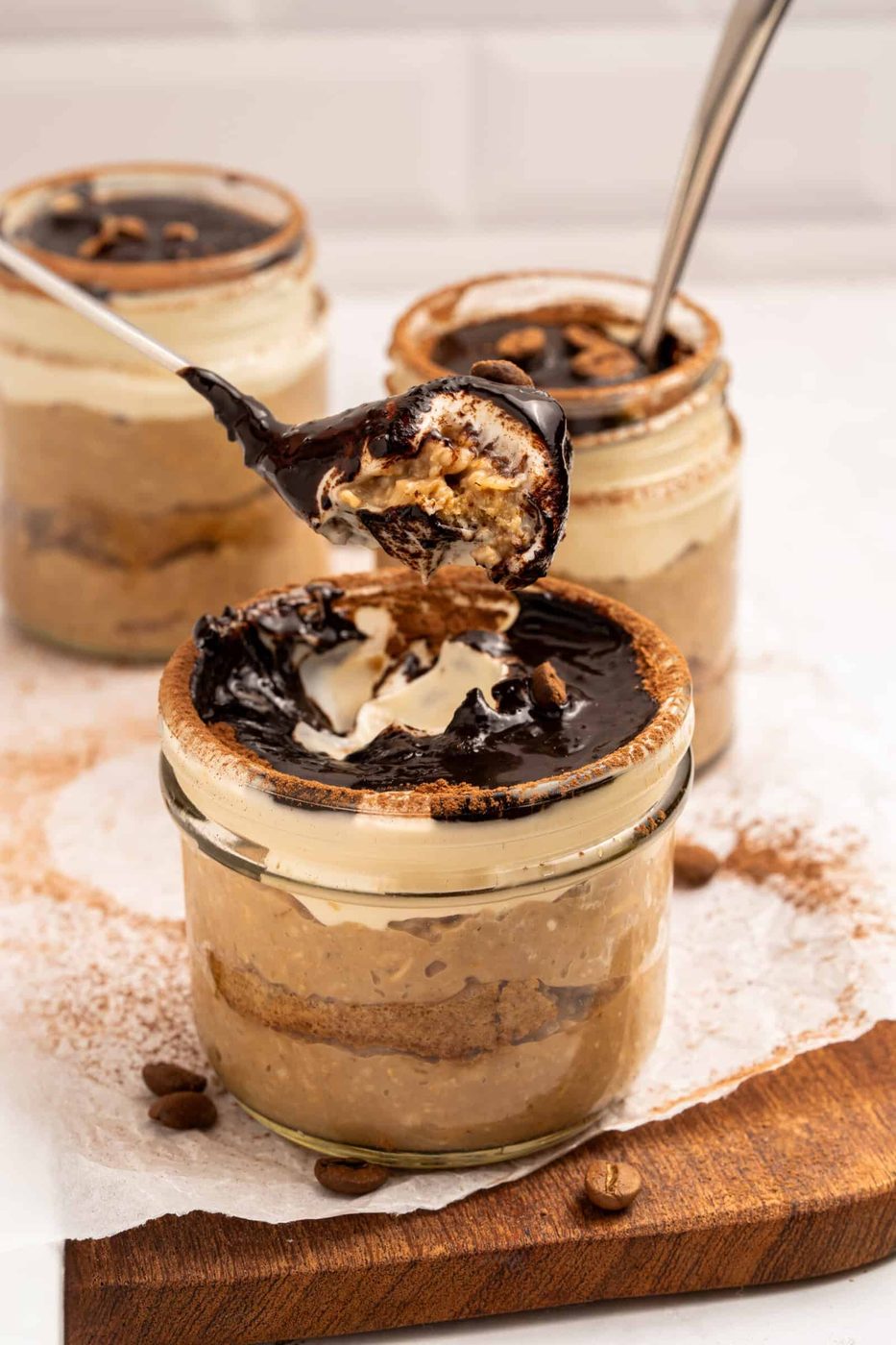 Three glass jars filled with layered tiramisu are topped with chocolate sauce and whipped cream. A spoon is lifting a portion from the front jar, revealing the creamy, layered inside. The jars sit on a wooden board with coffee beans.