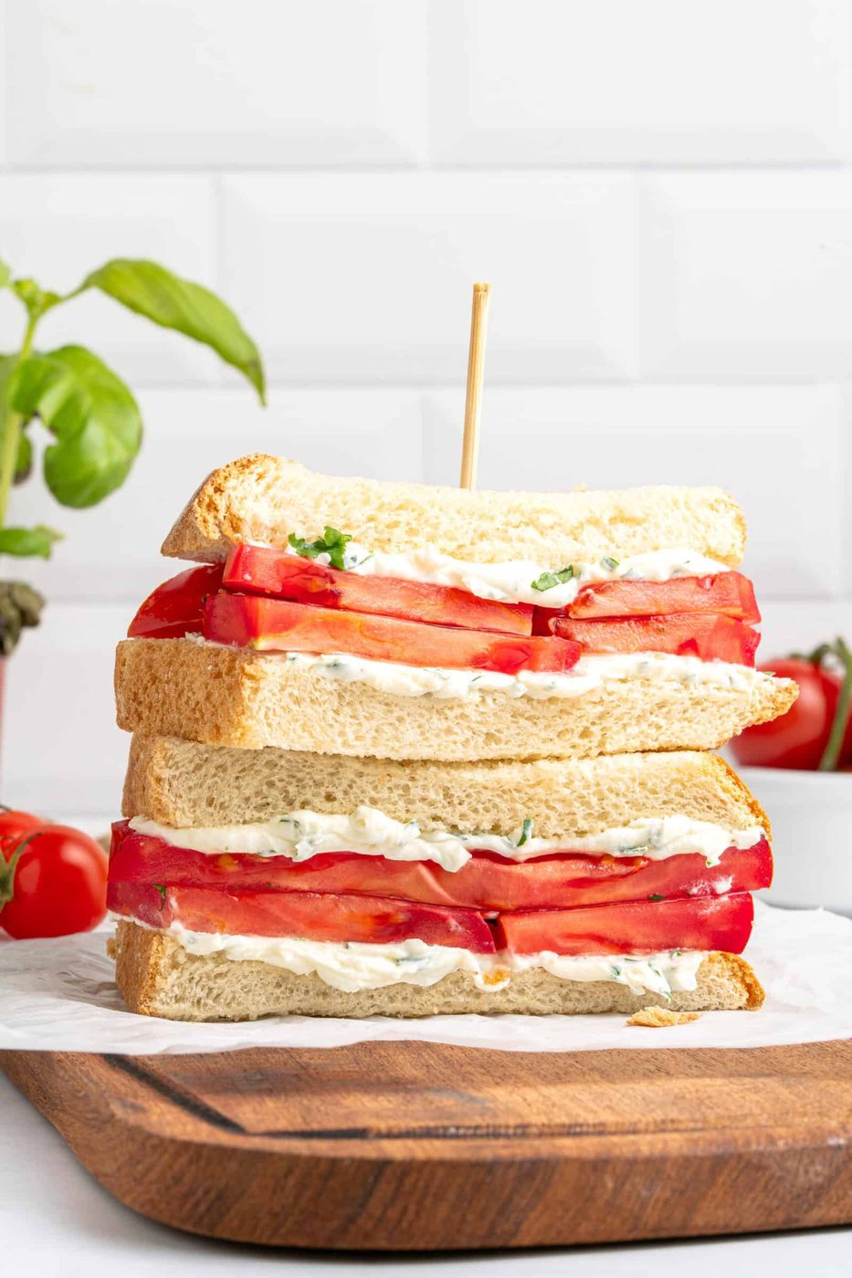 A stack of two tomato sandwiches with mayonnaise and herbs, made with thick slices of white bread, is shown on a wooden board. A skewer holds the sandwiches together. Fresh tomatoes and basil are in the background.