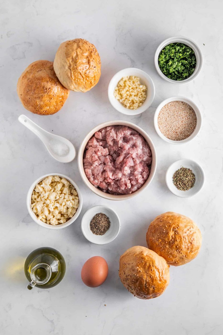 Ingredients for a recipe arranged on a marble surface include minced meat, chopped garlic, parsley, breadcrumbs, chopped onion, salt, pepper, whole egg, olive oil in a bottle, and four round buns.