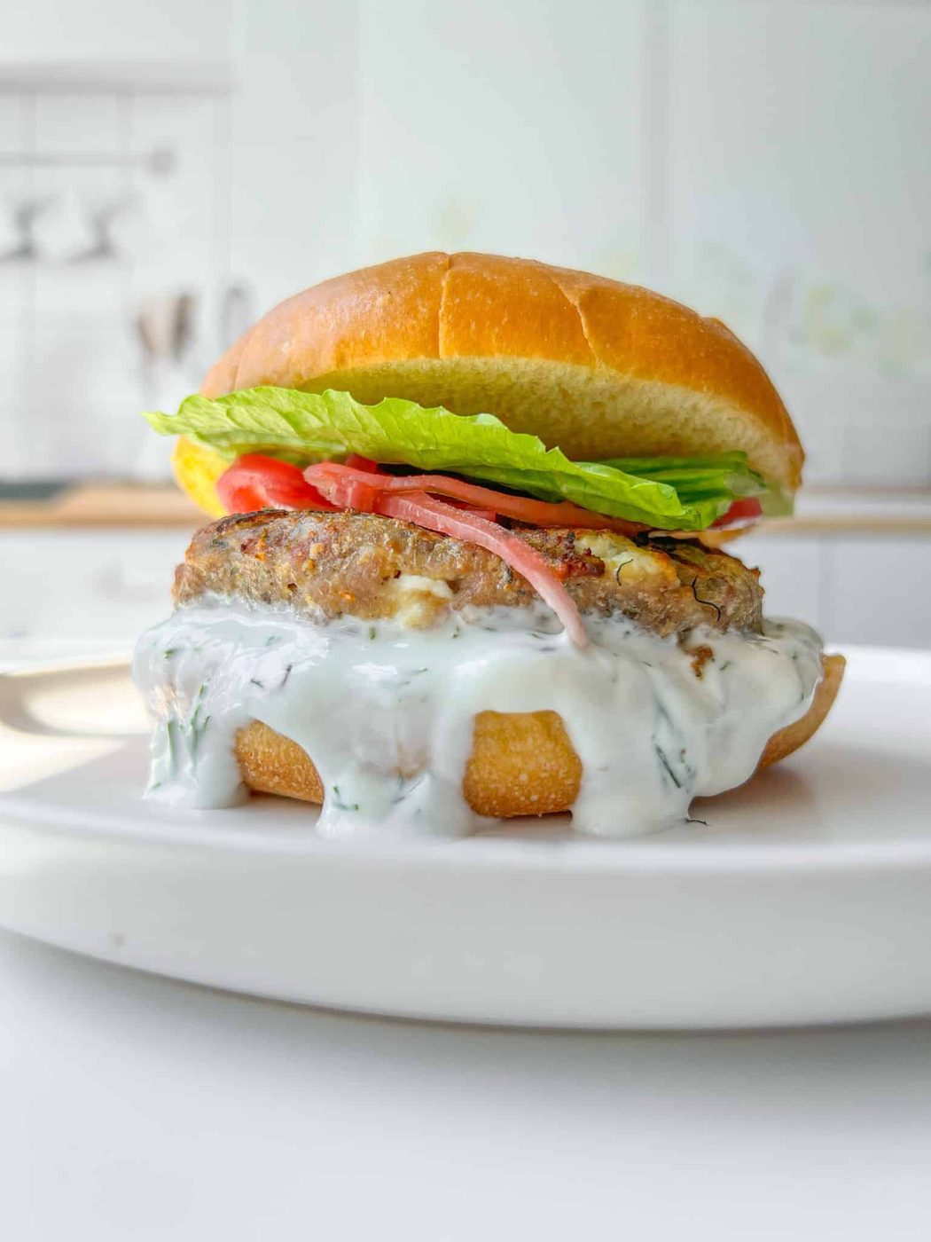 A close-up of a burger on a white plate. The burger features a creamy white sauce dripping down, a patty, tomato slices, and fresh lettuce, all sandwiched in a toasted bun. The background is a blurred kitchen setting.