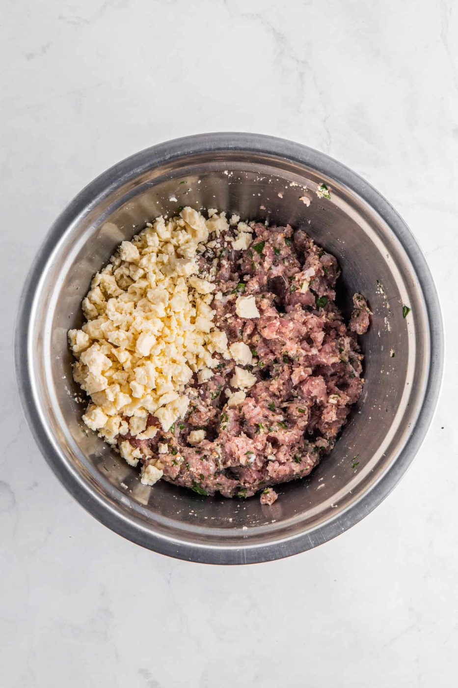 A metal bowl contains a mixture of ground meat with diced herbs on one side and crumbled cheese on the other, set against a white marble background.