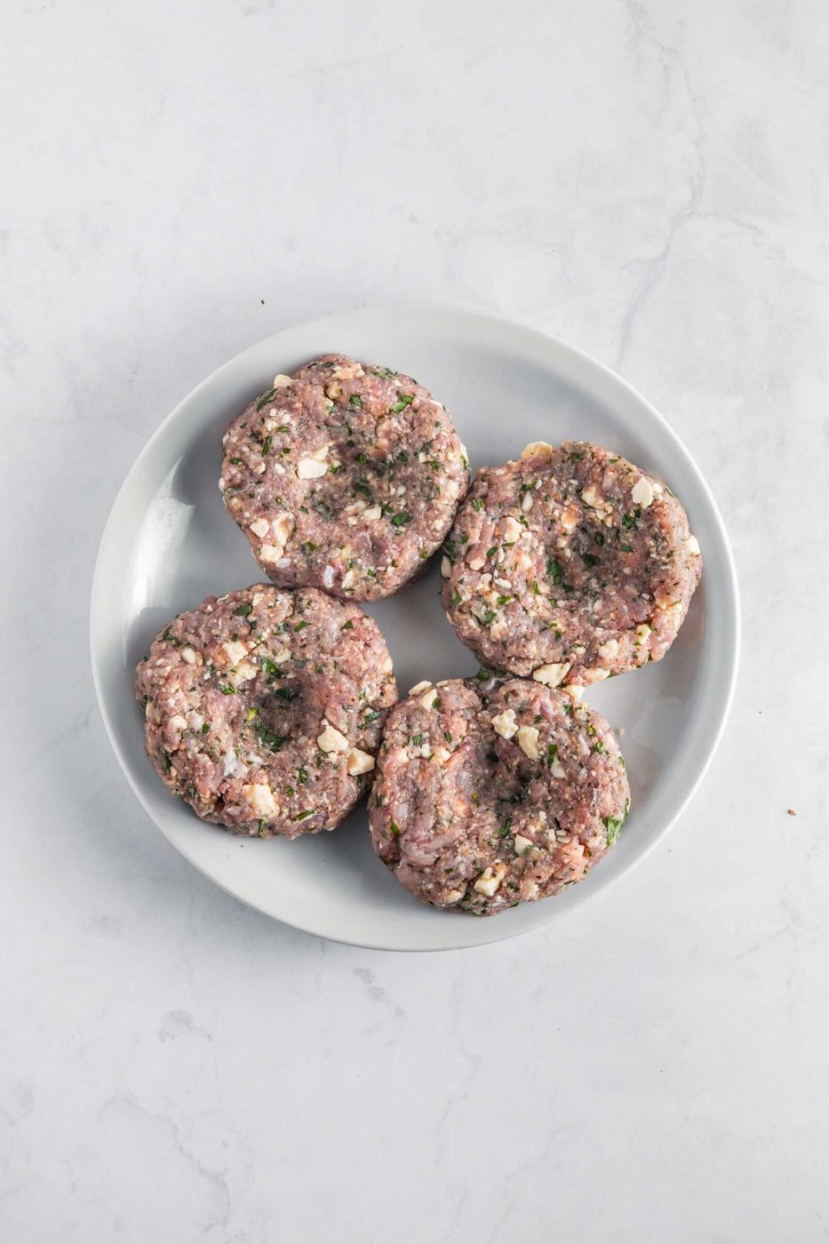Four uncooked burger patties made from ground meat mixed with herbs and chunks of cheese, placed on a white plate atop a light marble surface.