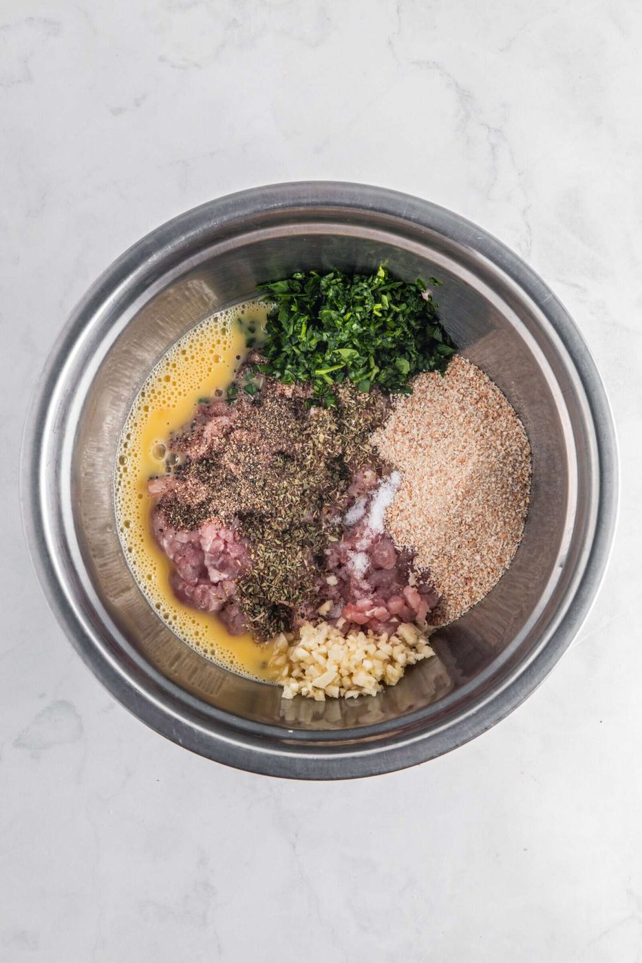 A metal bowl on a marble surface contains raw ground meat, breadcrumbs, chopped parsley, minced garlic, spices, and beaten eggs, all separated into sections, ready to be mixed for a recipe.