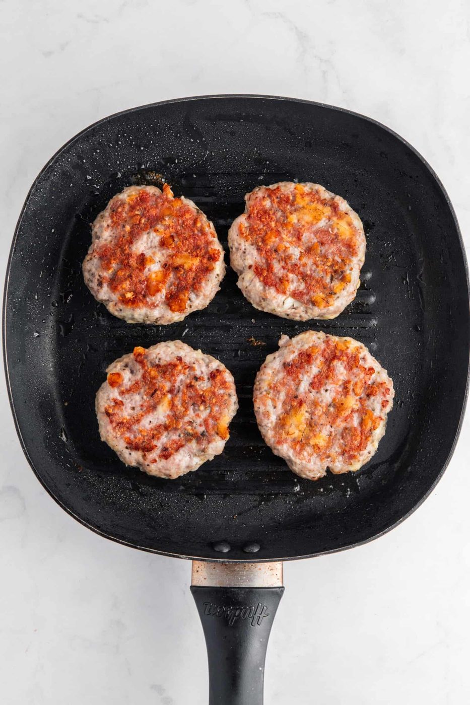 Four seasoned burger patties cooking on a black grill pan. The patties have a slightly charred texture with visible spices, and the pan has a handle facing downward. The background is a marble countertop.