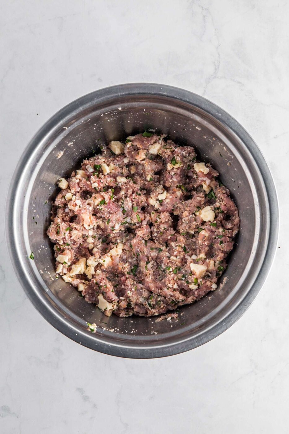 A metal bowl containing a mixture of minced meat with visible herbs and small pieces of onion, set on a light gray marbled surface.
