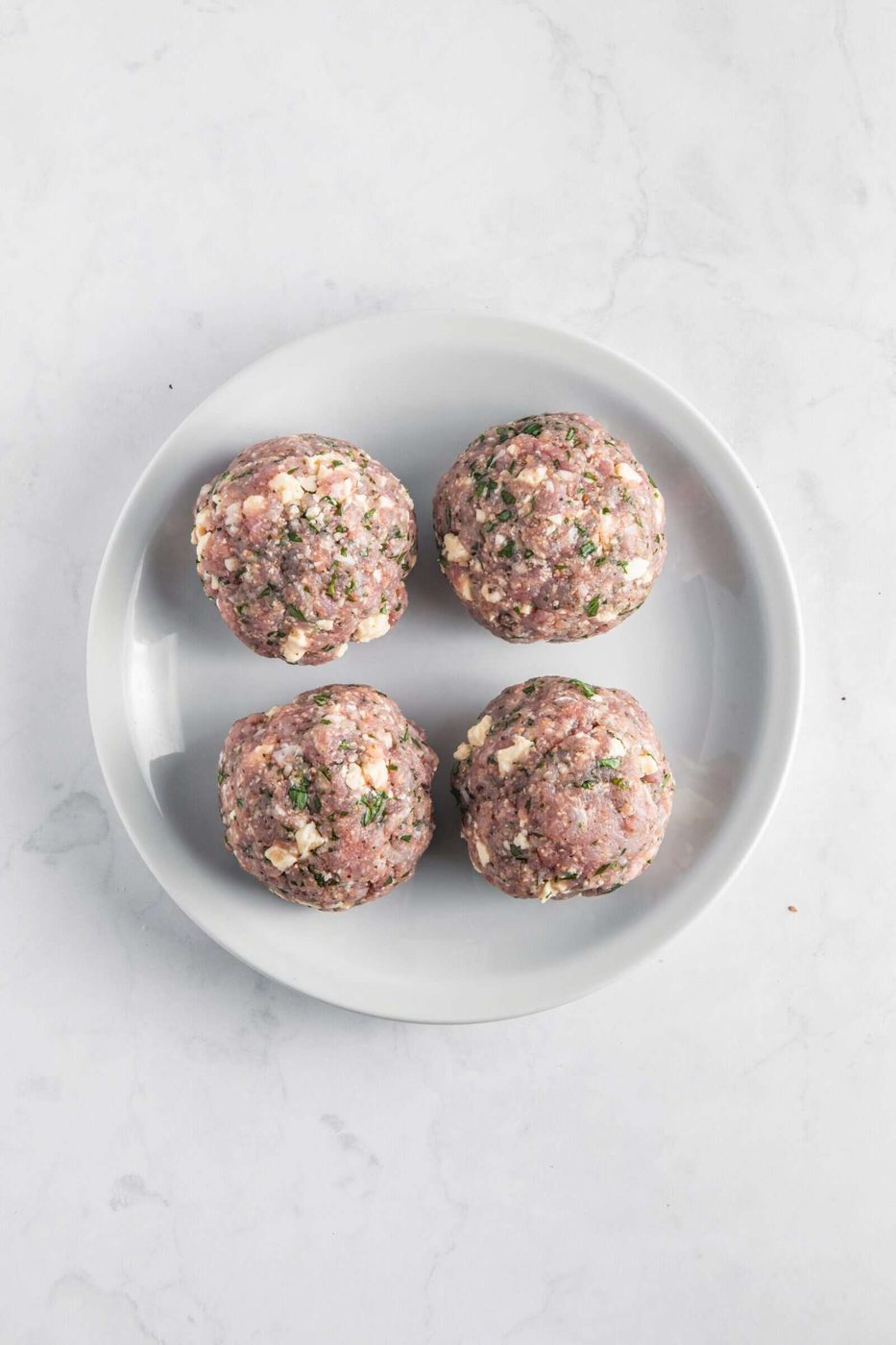 Four uncooked meatballs on a white plate against a light gray marble background. The meatballs appear to be seasoned with herbs and small pieces of chopped nuts or similar ingredients.
