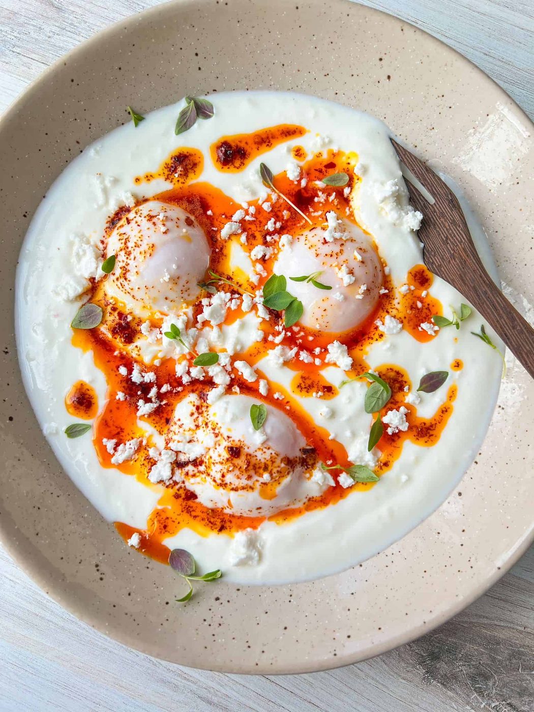 A dish featuring poached eggs on a bed of yogurt, drizzled with spicy red oil, garnished with crumbled feta cheese and fresh herbs. A wooden spoon rests on the beige plate.