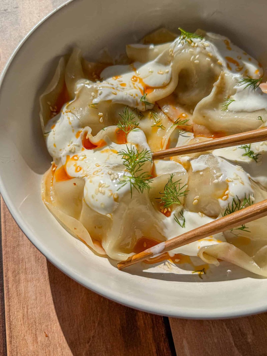 A bowl of Blanket Dumplings topped with creamy white sauce, chili oil, sesame seeds, and fresh dill, with a pair of wooden chopsticks resting on the side. Warm sunlight illuminates the dish.