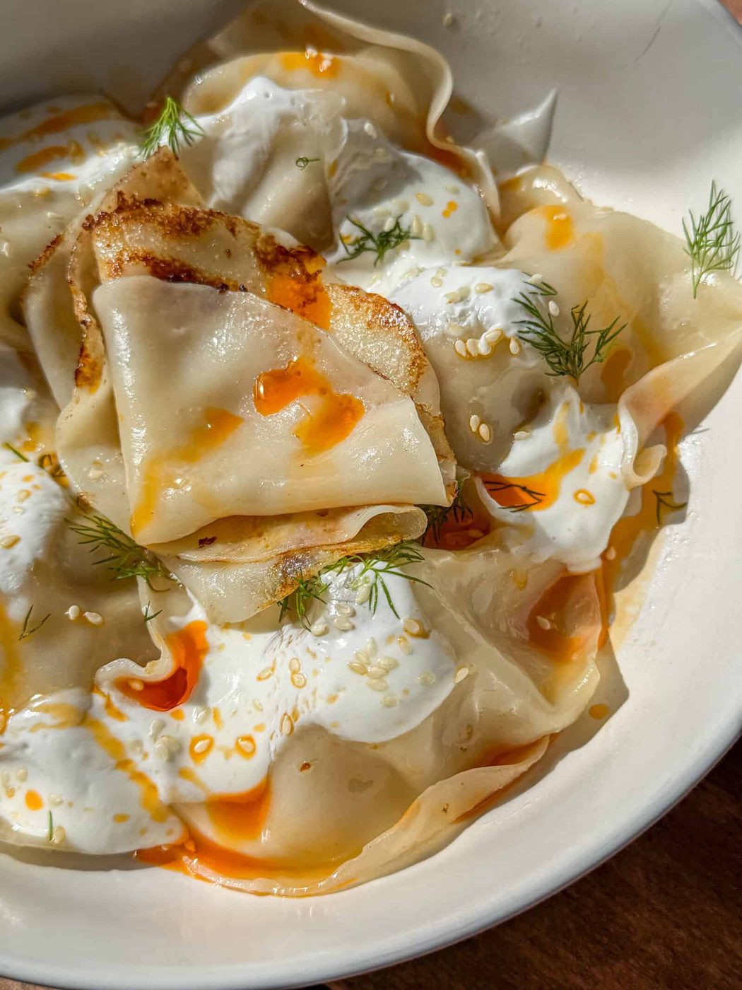 A close-up of Blanket Dumplings topped with creamy white sauce, orange chili oil, sesame seeds, and fresh dill, served in a white bowl.
