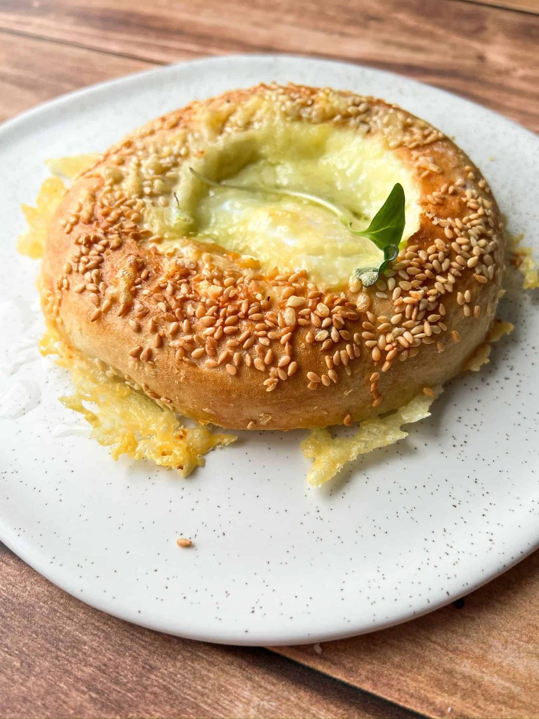 A round, baked bread topped with sesame seeds and melted cheese, garnished with a small green leaf, sits on a white speckled plate on a wooden surface.