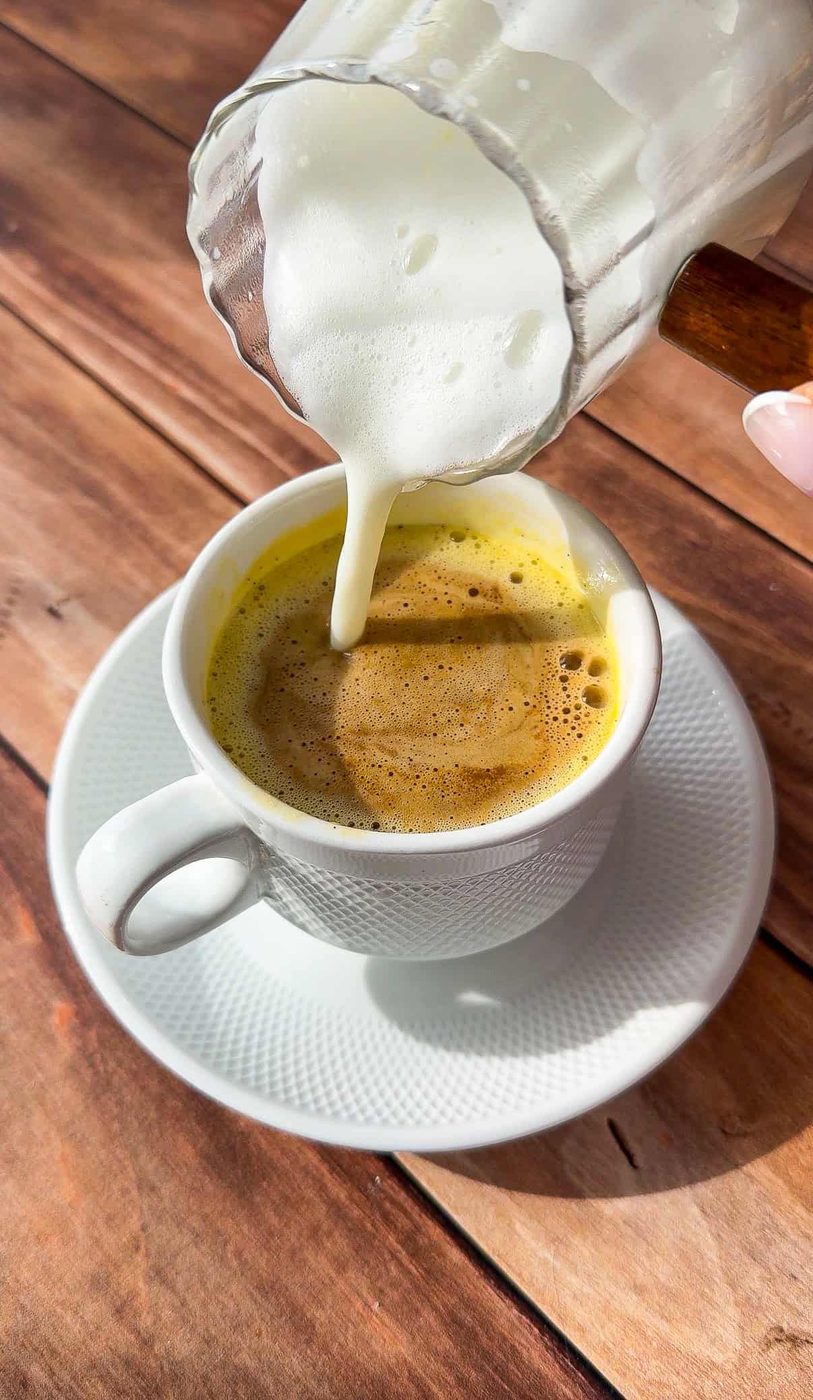 A hand pours steamed milk from a glass pitcher into a cup of Egg Coffee on a white saucer, set on a wooden table in natural sunlight.