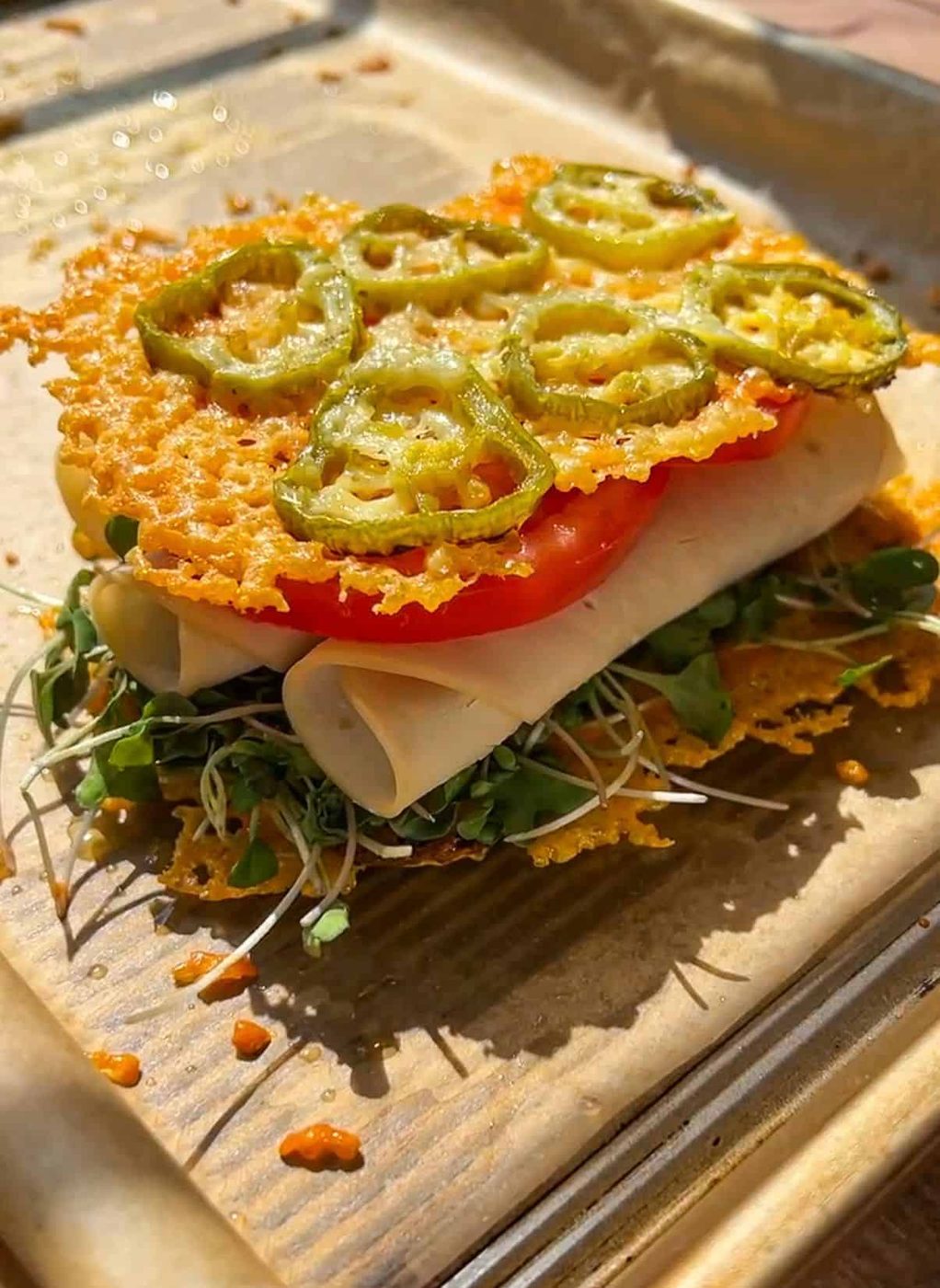 A sandwich with microgreens, rolled turkey slices, tomato, and crispy baked cheese topped with jalapeño slices, served on parchment paper.