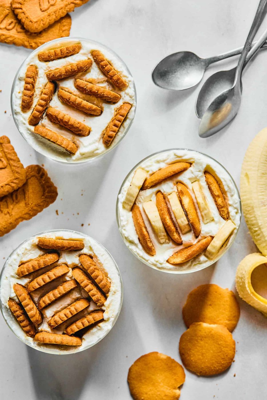 Three dessert cups topped with cookies and banana slices are arranged on a white surface, inspired by the Japanese Yogurt Cheesecake trend, surrounded by extra cookies, banana slices, and three metal spoons.