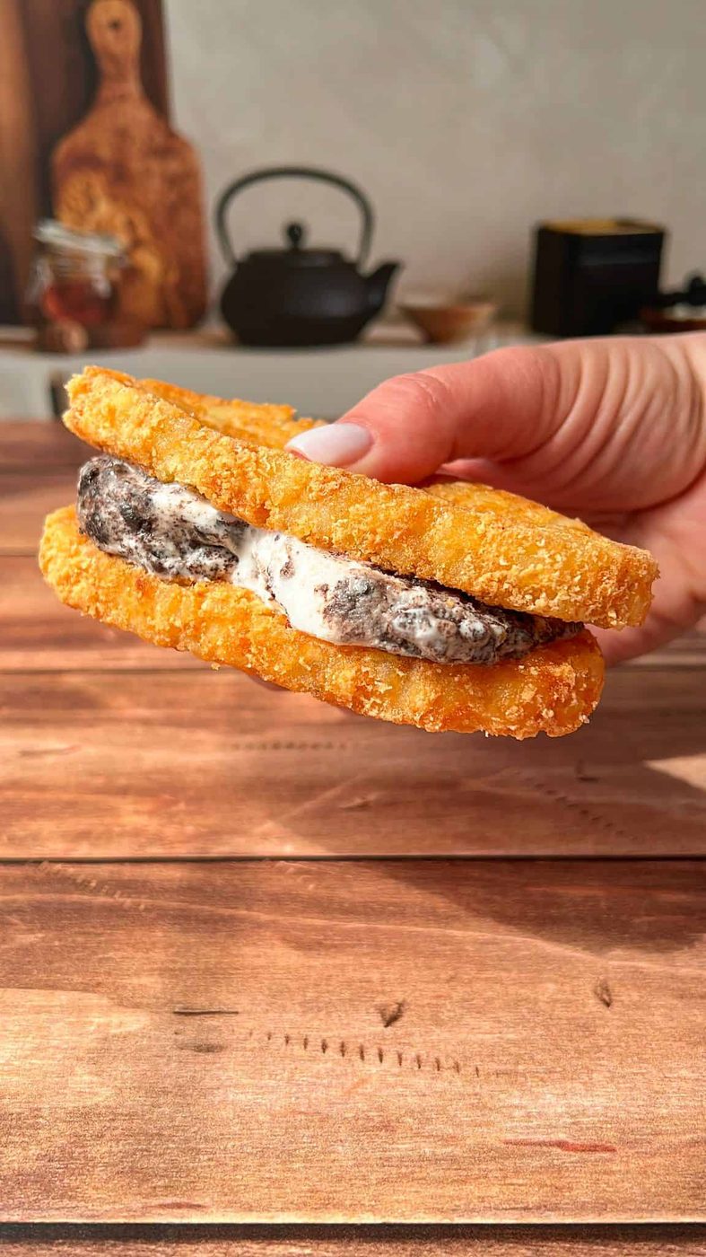 A hand holds a sandwich made with two crispy, breaded hash browns as buns, filled with cookies and cream ice cream, against a wooden table background with kitchen items blurred in the distance.
