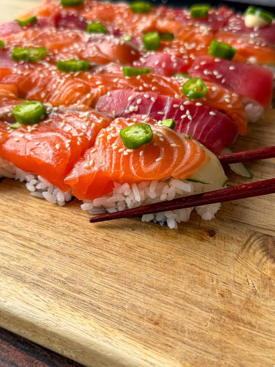 Close-up of assorted sushi topped with fresh slices of salmon and tuna, green wasabi dollops, and sesame seeds, arranged on a wooden board with a pair of chopsticks picking up a piece.