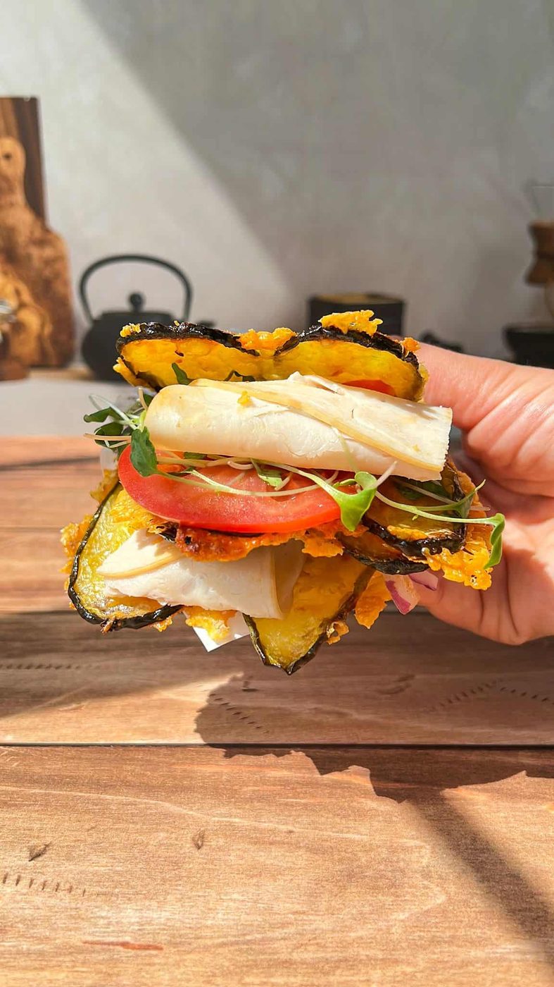 A hand holds a pickle sandwich made with crispy, golden plantain slices instead of bread, filled with tomato, cheese, greens, and white vegetable slices, all set on a wooden table in bright sunlight.