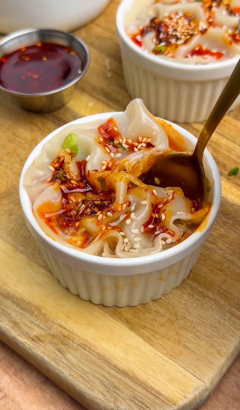 A white ramekin filled with dumplings topped with chili oil, sesame seeds, and green onions sits on a wooden board. A spoon is scooping a dumpling, and a small dish of chili oil is nearby.