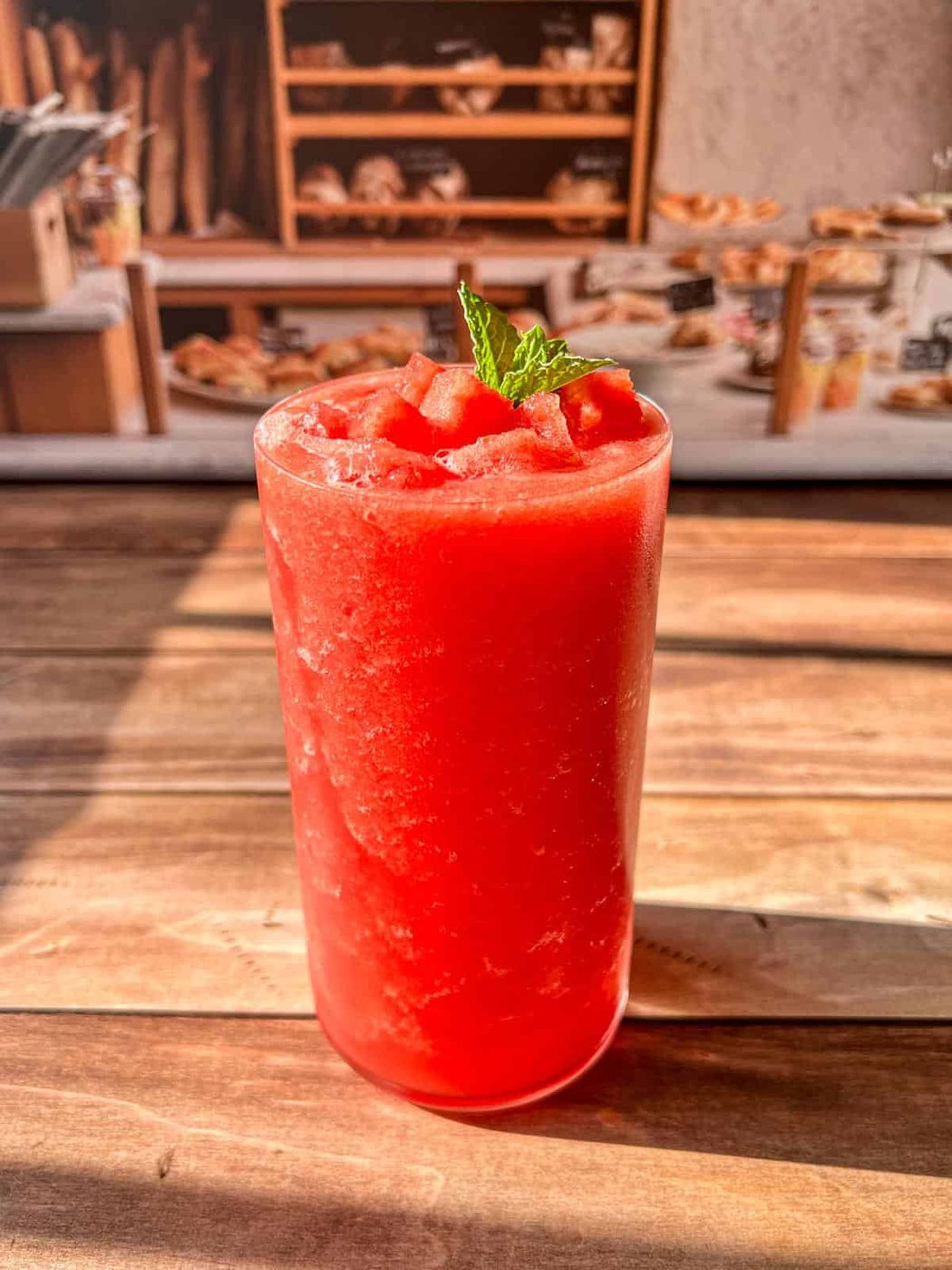 A tall glass filled with bright red watermelon smoothie, topped with small watermelon pieces and a mint sprig, sits on a wooden table. The background shows a blurred bakery display with shelves of bread and pastries.
