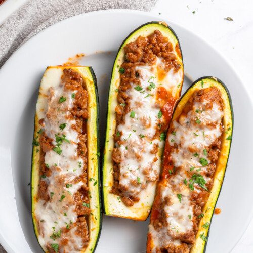 Three Stuffed Zucchini Boats filled with a savory ground meat and tomato mixture, topped with melted cheese and chopped herbs, served on a white plate.