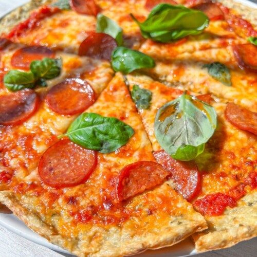 A close-up of a Low Carb Pizza topped with pepperoni and fresh basil leaves, sliced into pieces and served on a white plate. The melted cheese and tomato sauce are visible in bright, natural sunlight.