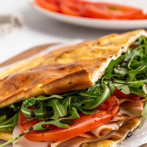A freshly made sandwich on a wooden board, filled with sliced turkey, fresh arugula, and tomato slices. The sandwich is on ciabatta bread, slightly toasted. A white plate with additional tomato slices is blurred in the background.