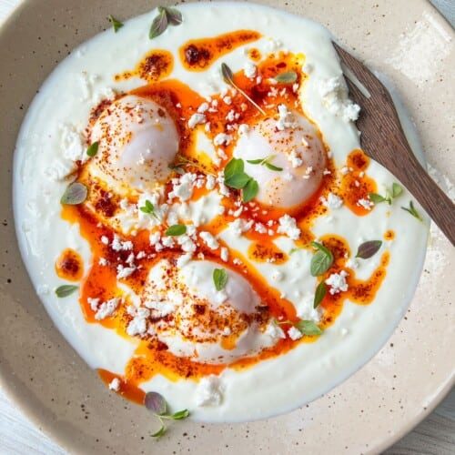 A dish featuring poached eggs on a bed of yogurt, drizzled with spicy red oil, garnished with crumbled feta cheese and fresh herbs. A wooden spoon rests on the beige plate.