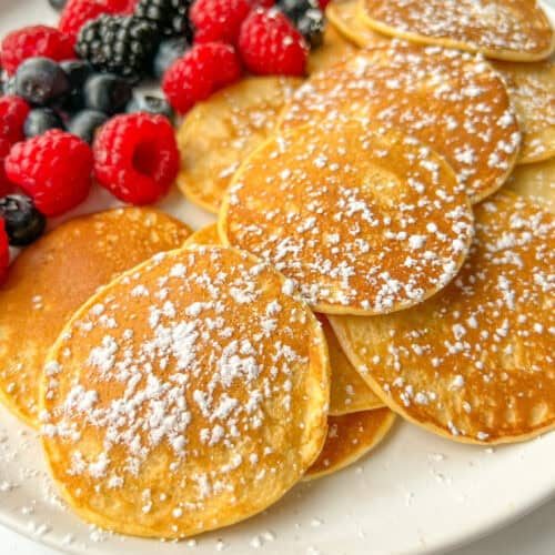 A plate of mini cottage cheese pancakes sprinkled with powdered sugar, served with fresh raspberries, blackberries, and blueberries.