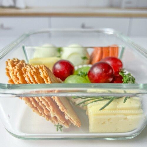 A glass container filled with sliced crackers, cheese, green and red grapes, carrot sticks, and cucumber slices creates the perfect Adult Lunchable, placed on a white surface with a blurred kitchen background.