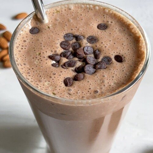 A glass of chocolate smoothie topped with chocolate chips, served with a metal straw. Some whole almonds are scattered in the background on a light surface.