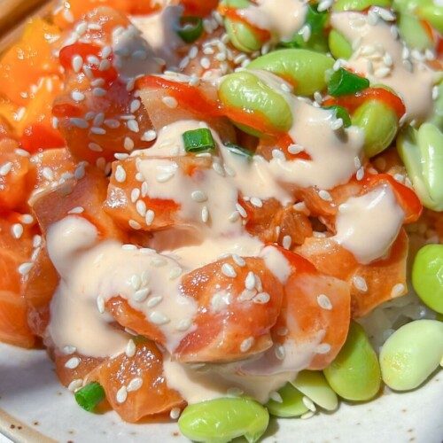 A close-up of a Spicy Salmon Poke Bowl featuring diced raw salmon, edamame beans, sesame seeds, chopped green onions, and creamy dressing, served on a white plate with chopsticks.