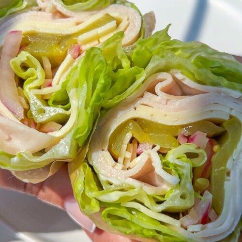 Close-up of two hands holding a Lettuce Wrap made with layers of lettuce, sliced turkey, cheese, pickles, and chopped vegetables—no bread—in front of a light background.