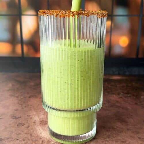 A tall, ridged glass is being filled with a smooth green smoothie. The rim of the glass is coated with a spice mix. The background is softly blurred with warm, golden hues, suggesting evening light through a window.