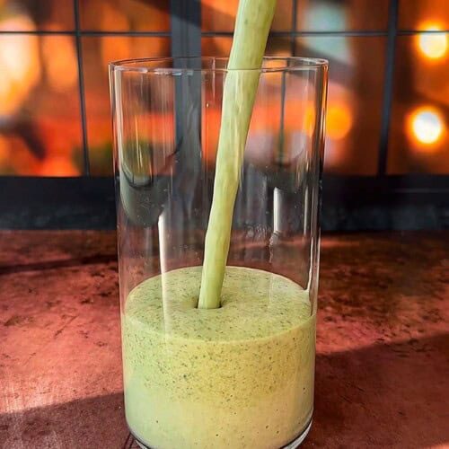 A thick, green smoothie is being poured into a clear glass. The background features a warm, out-of-focus display of glowing lights, creating a cozy ambiance. The scene is set on a wooden surface.