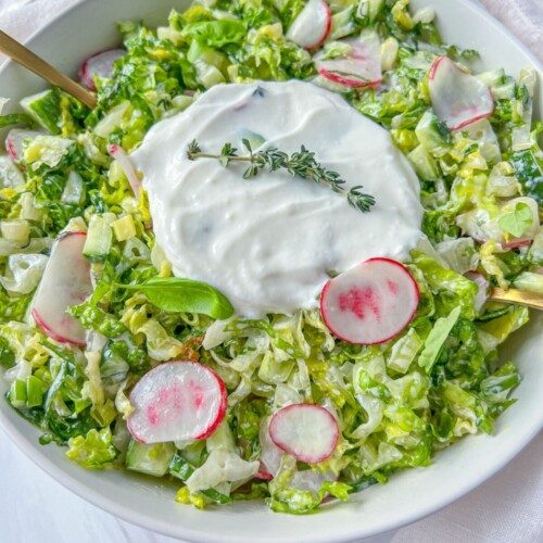 A bowl of fresh chopped salad inspired by Bulgarian Salad, with lettuce, sliced radishes, and cucumbers, topped with creamy white dressing and garnished with sprigs of fresh thyme. Two gold utensils rest in the bowl.