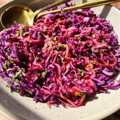 A bowl of vibrant red cabbage slaw with finely shredded purple cabbage, carrots, and herbs, topped with a light vinaigrette. A gold fork and spoon rest beside the dish on a wooden table.